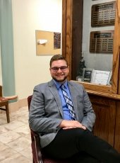 A young man in a tie smiles with a bookshelf behind him in the MTU College of Business