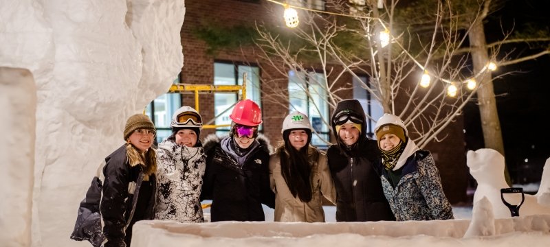 six girls in hard harts and winter hats and coats smile underneath outdoor lights framed by their snow statue in front of MTU's administration building in winter.