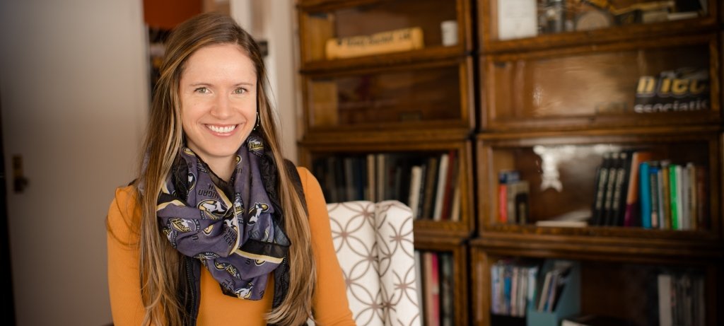 A woman in a Michigan Tech Husky scarf smiles sitting in a chair by a bookshelf.