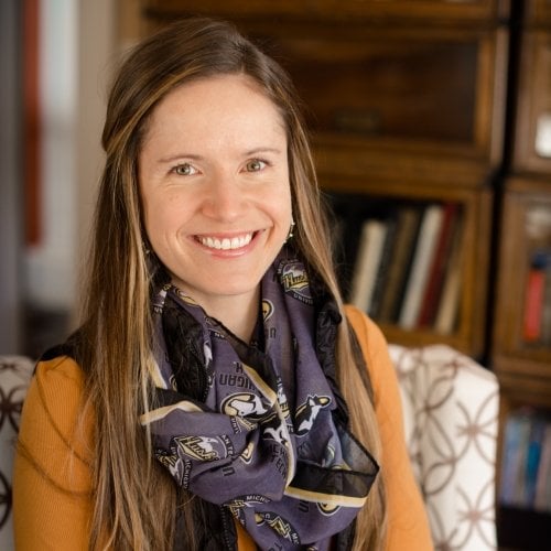 A smiling woman wearing a Michigan Tech Husky scarf and earrings sits on a chair with a bookcase behind her.