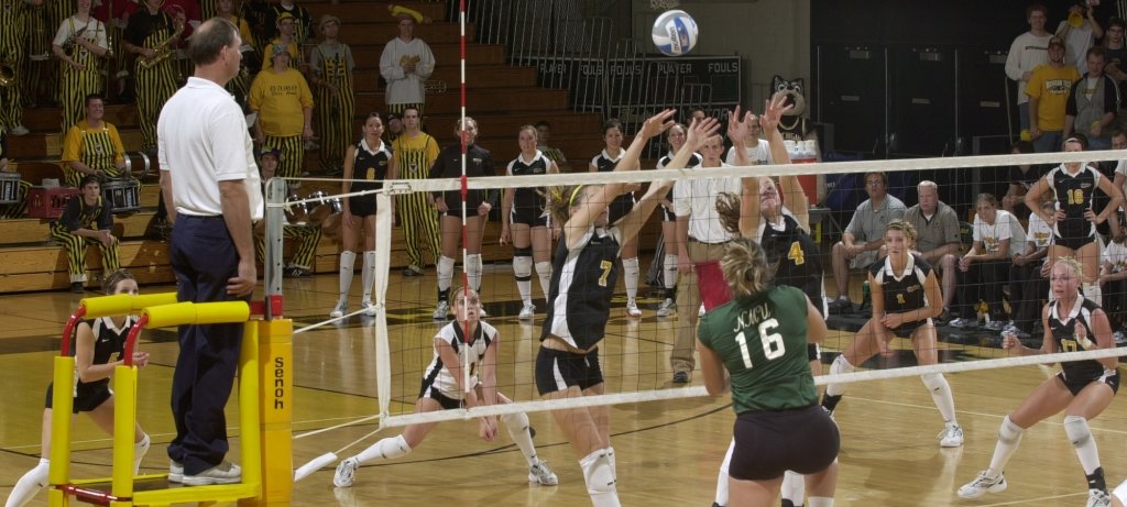 A college womens volleyball game with number 7 and 4 going for the ball as No. 16 tries to block it in a gym with spectators in black and gold stripes and a ref to the left.