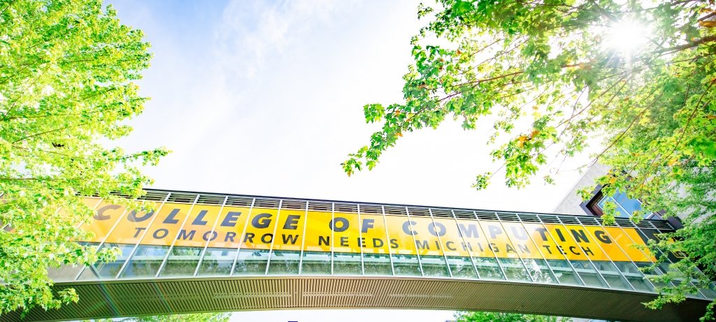 College of Computing Tomorrow Needs Michigan Tech reads the gold banner on a glass walkway with blue sky and green trees arching overhead.