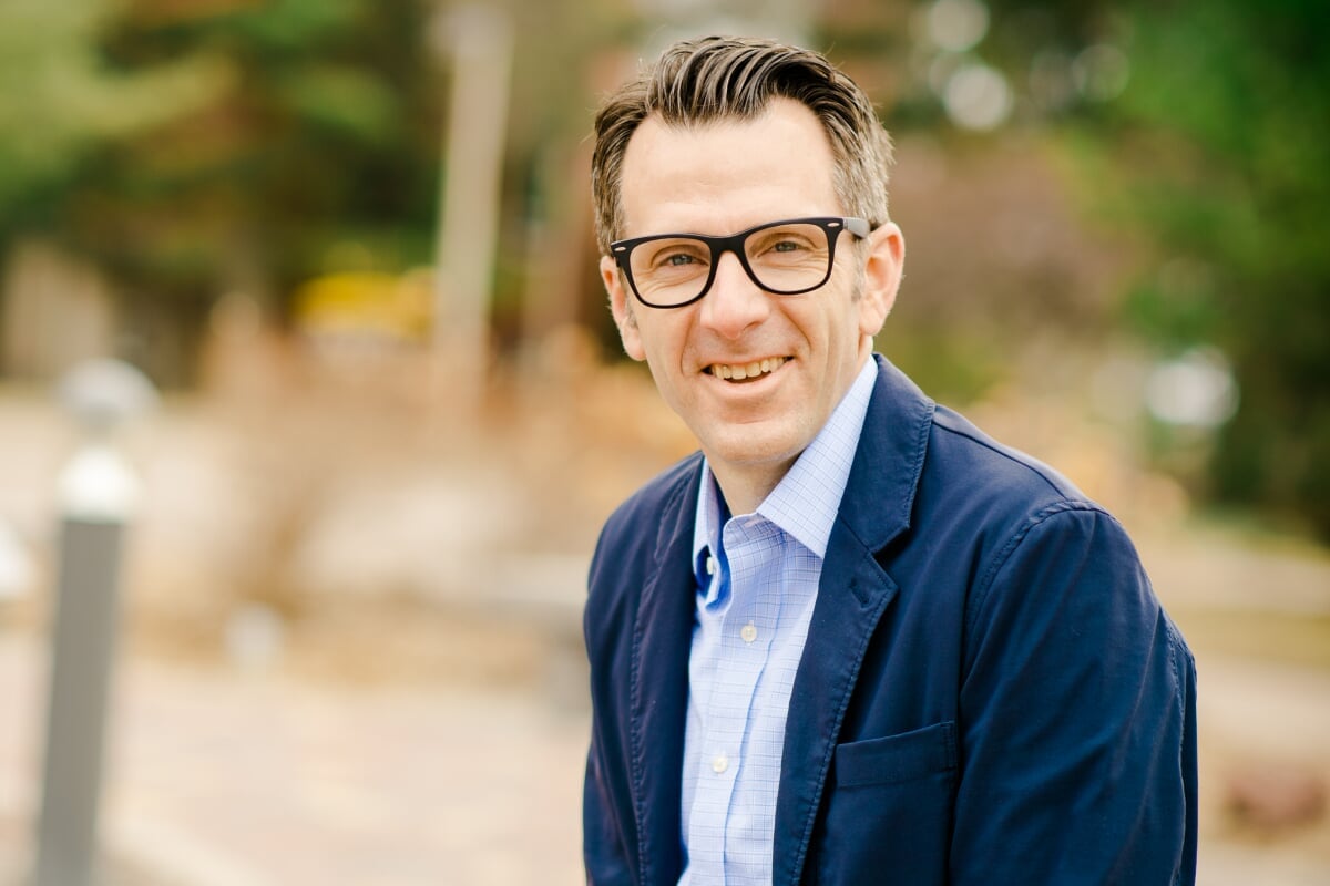 A man with glasses smiles at the camera with trees behind him on campus.