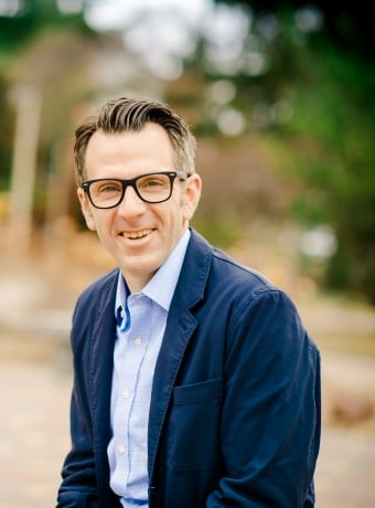 A man with glasses smiles at the camera with trees behind him on campus.