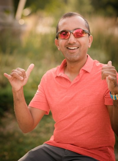 A faculty member in shiny sunnies signals 'I love you" with trees in soft focus in the background on the Michigan Tech Campus.