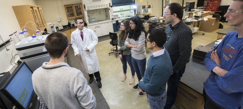 A researcher in a lab coat talks to six students in his lab at Michigan Tech.