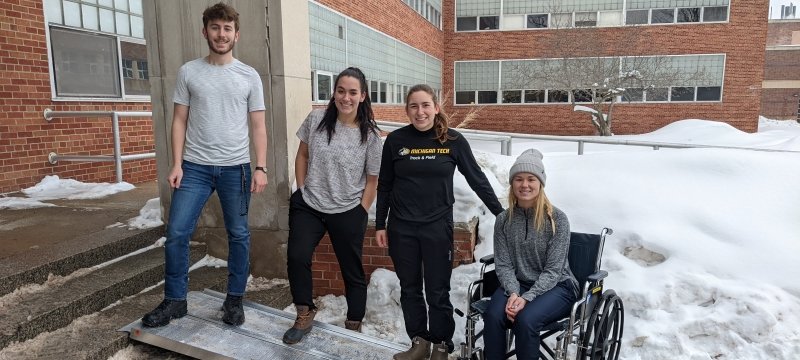 four students stand in a row on a wheelchair ramp with the final student in a wheelchair on a snowy day outside Dillman Hall on the MTU campus showing their invention.