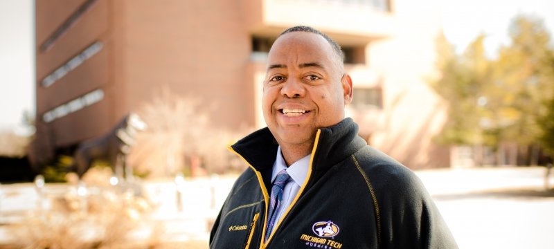 A man wearing a Michigan Tech Huskies jacket smiles on campus with soft focus trees and brick building in the background.