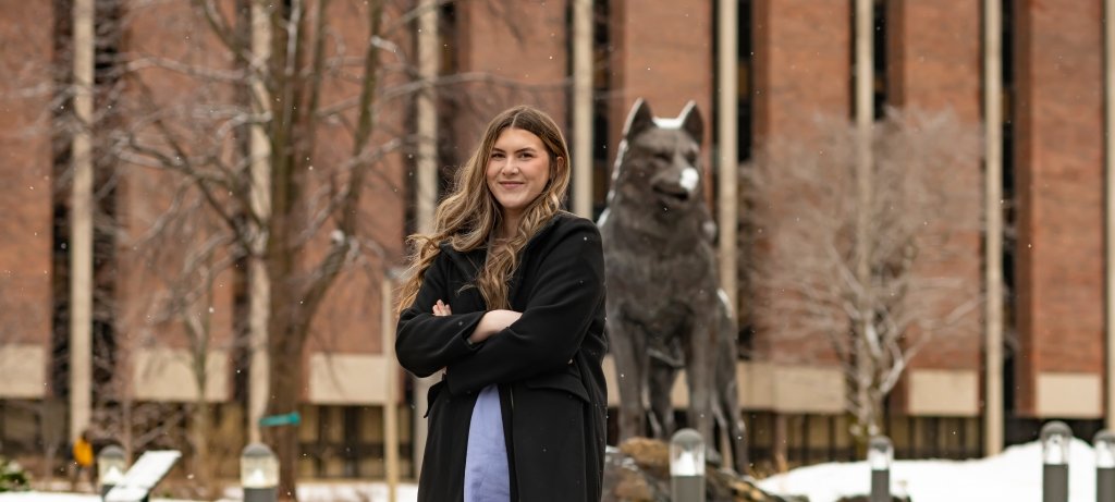 A young woman stands in Husky Plaza at Michigan Tech in front of the Husky statue. She is student speaker for springcommencement 2022.