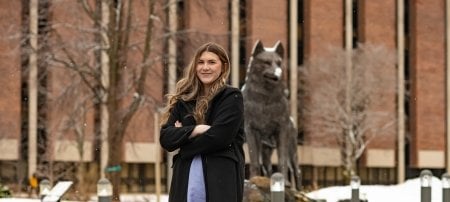 A young woman stands in Husky Plaza at Michigan Tech in front of the Husky statue. She is student speaker for springcommencement 2022.