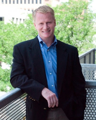 A man stands with green trees behind him on a balcony railing; John Bacon is spring 2022 commencement speaker.