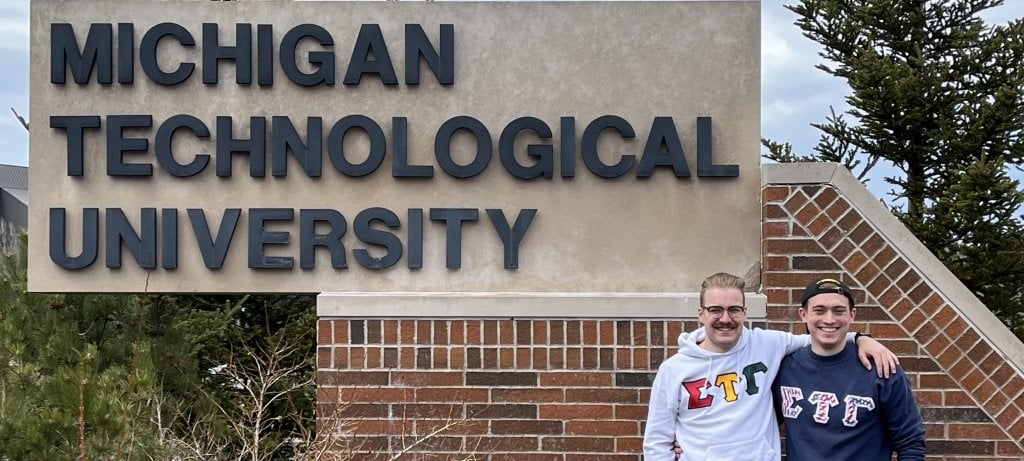 Two students who went to Michigan Tech together stand in front of the University sign outside the year they came to MTU.