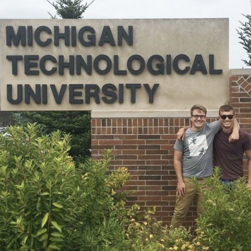 In front of the Michigan Technological University sign, two childhood friends and grads on the day they came to MTU.