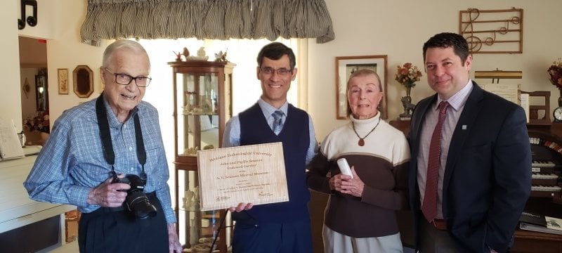 Four people in a living with notes on a wall and a museum benefactor holding a plaque.