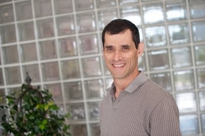 A man who is curator of a mineral museum smiles at the camera with clear glass background behind him and a green plant.