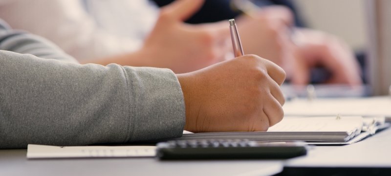 Close-up picture of a student writing in a notebook with a mobile device nearby.