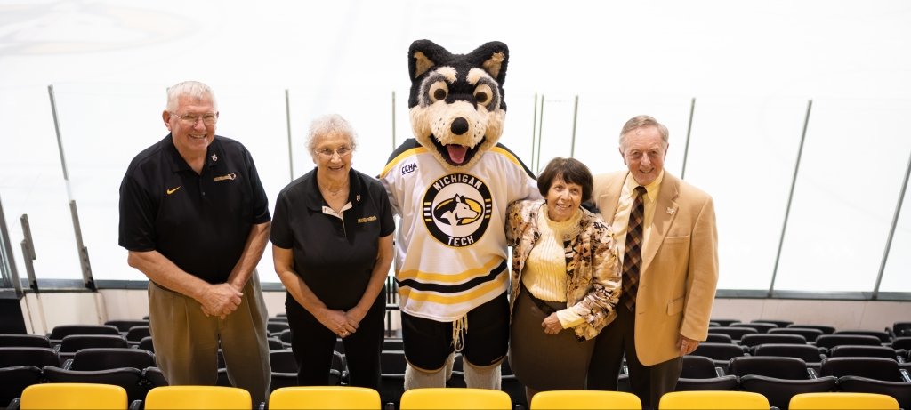 Two couples flank Blizzard T. Husky in Michigan Tech's MacInnes Student Ice Arena. Mascot supporters are to the left, and Tech's original mascots are to the right.