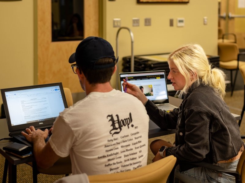 A student with a laptop helps a fellow student at a laptop with their resume in the common space at Michigan Tech Career Services. They're getting ready for in-person Career Fair.