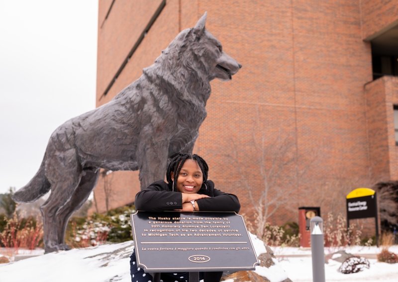 A young woman leans on a sign by the Husky statue outside in Michgan Tech's Husky Plaza, smiling as she prepares to graduate.