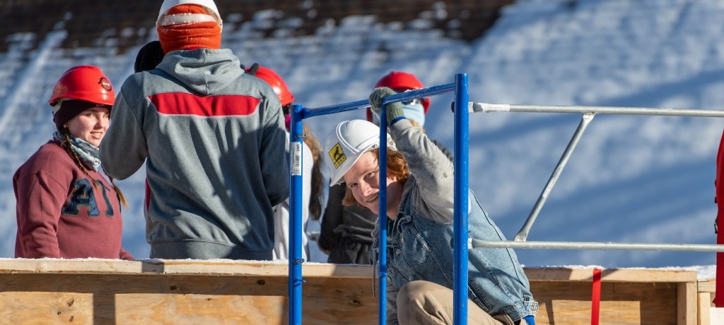 Students at Michigan Tech on a roof-high snow statue scaffold smile on campus as they prep for Michigan Tech Winter Carnival.