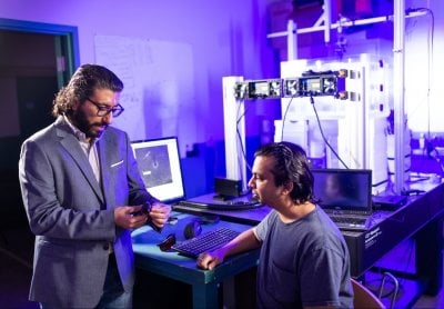 Two researchers stand near a tank and computer monitoring station in the ultraviolet purple-lit fluid dynamics and active matter lab.