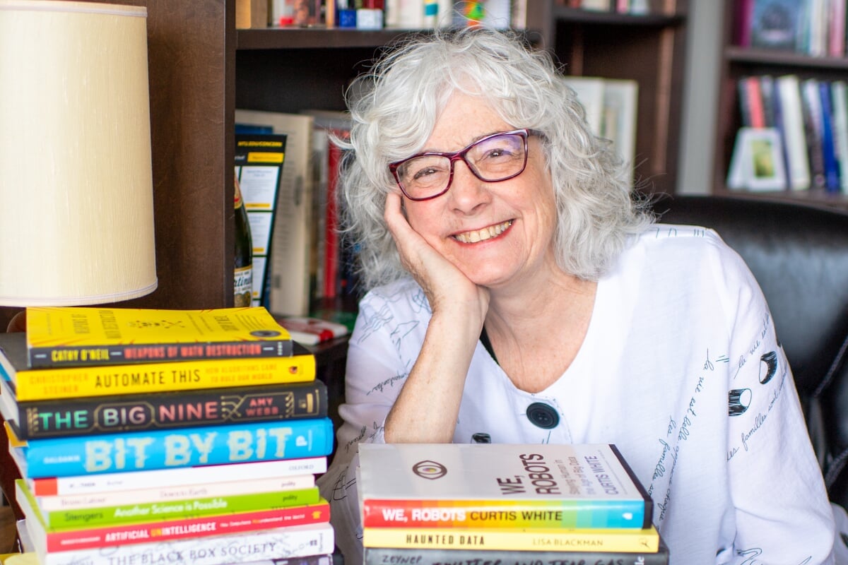 Professor Jennifer Slack smiles in her office as she sits with a pile of books related to new media and algorithmic culture in the Michigan Tech Institute of Policy, Ethics, and Culture.