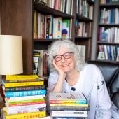 Professor Jennifer Slack smiles in her office as she sits with a pile of books related to new media and algorithmic culture in the Michigan Tech Institute of Policy, Ethics, and Culture.