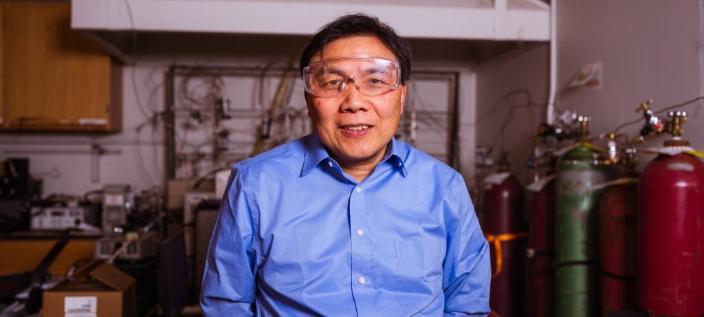 A Michigan Tech researcher who discovered a new fuel cell stands in his lab with fuel tanks in the background. He's wearing safety glasses.