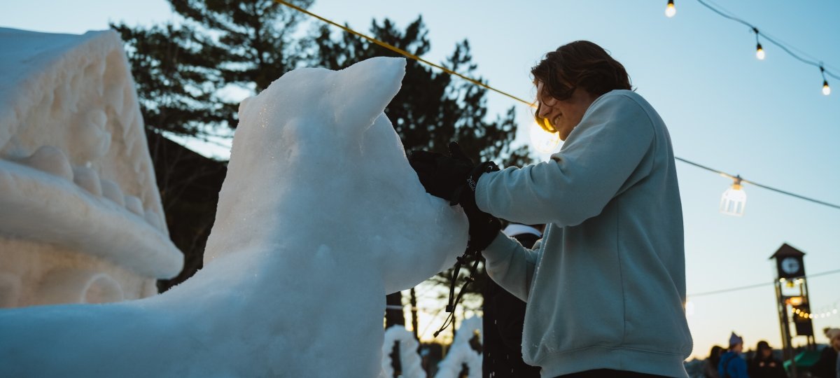 A student stands in front of an ice sculpture of a cow during Michigan Tech Winter Carnival. The campus clocktower and string lights are in the background.