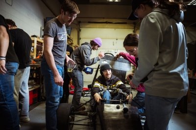 Michigan Tech's formula SAE Enterprise team is one of several in the collegiate automotive design programs offered at the university, in which teams work together to build vehicles with greater capabilities and effiencies. A team member is in the vehicle and fellow Huskies are clustered around him, making adjustments to the engine and body of the vehicle. Others at the side do research at a workbench.