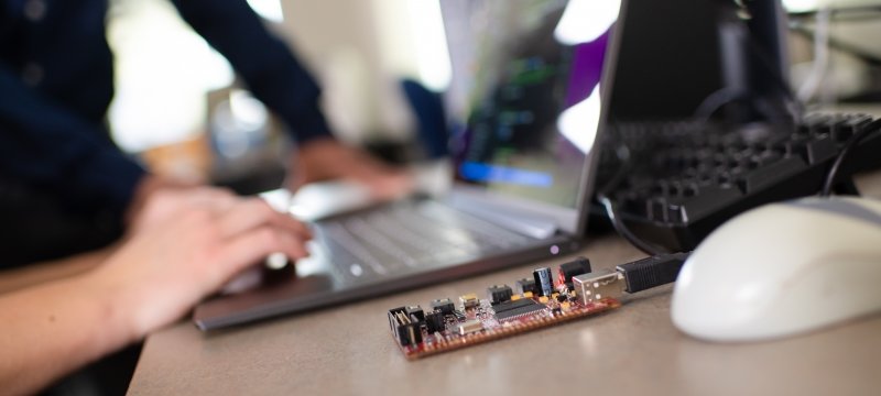 A computer in Bo Chen's cybersecurity lab at Michigan Tech with a circuit board and mouse in the foreground as a researcher's hand is shown navigating the cursor.