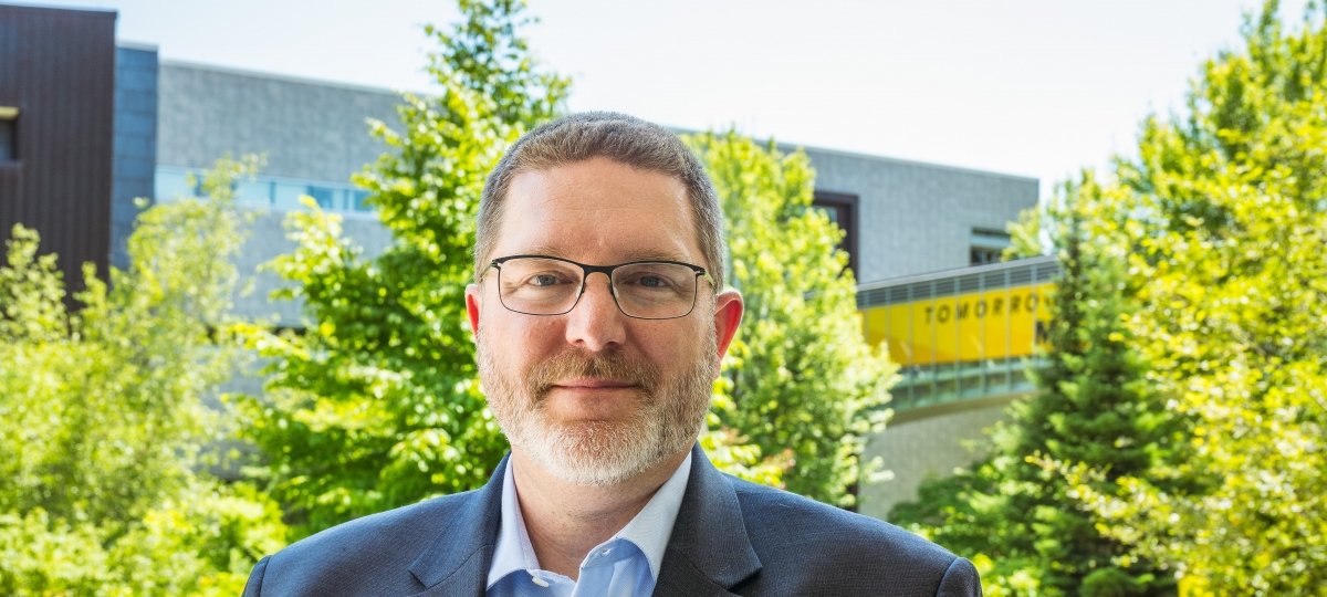 Andrew Barnard on campus with the Library/Rekhi Hall bridge in the background.