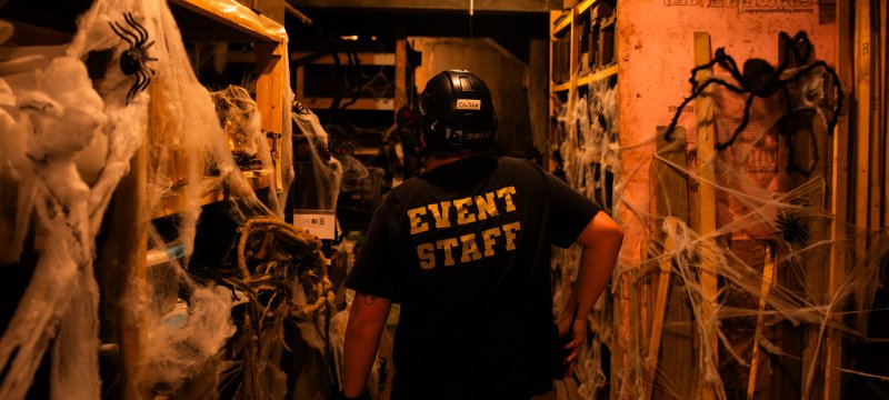 Person wearing an Event Staff shirt walking through a dark area with shelves covered in fake spiders and webs.