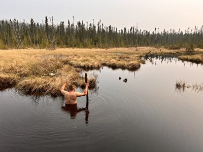A man with a staff waist-deep in water with his back toward the camera and arms up wades toward bones in a marshy bog on a mat of grass in a clearing.