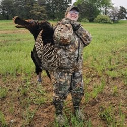 Blissick stands in an open field wearing camouflage hunting gear, hefting a freshly shot turkey over her shoulder.