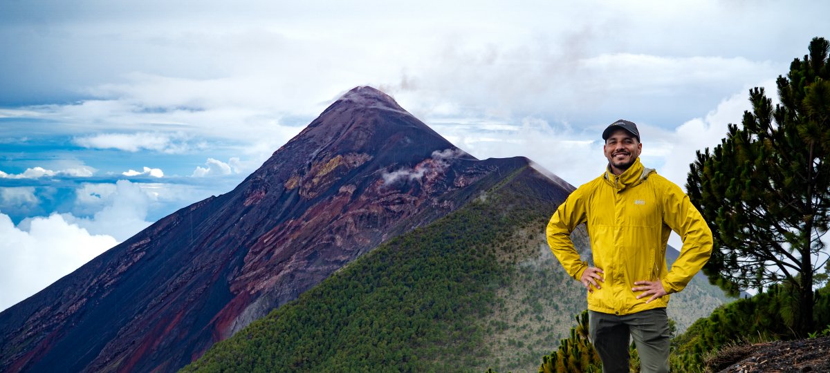 Gustavo Bejar-Lopez has been interested in volcanoes since he was a child. Now, he's working to help better prepare people for volcanic hazards and to mentor a future generation of volcanologists. (Photo courtesy Gustavo Bejar-Lopez) Man standing hands on hip in a yellow jacket, smiling in front of Peak of Volcan de Fuego, a volcano, in the background.
