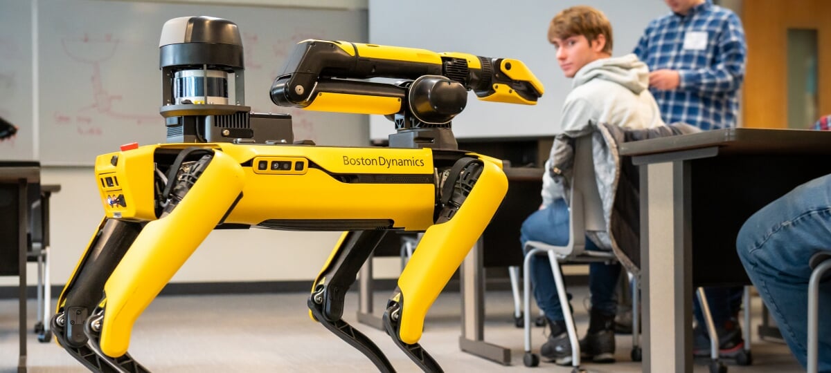 Balto the robot dog runs through its paces for students and faculty in the Michigan Tech College of Computing. Student in the background at a desk turned around and looking at the yellow and black robot dog.