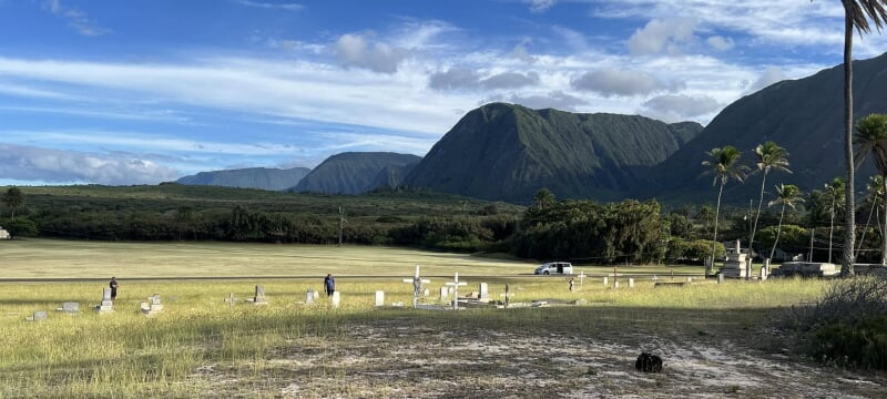 A couple people walking through a field with some grave markers with mountains in the distance.