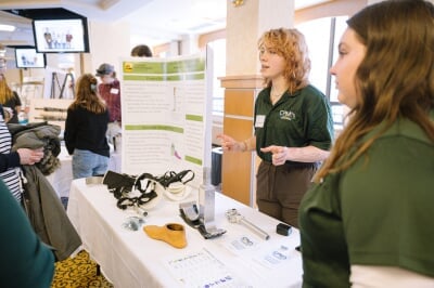 Student members of the Consumer Product Manufacturing Enterprise present on their team's work, wearing green polos that read 'CPM Enterprise.' On the table in front of them is a disassembled prosthetic leg and foot.