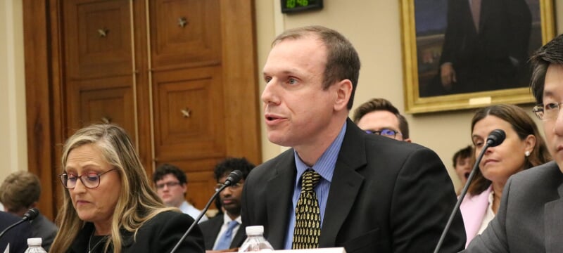 Stephen Techmann talks into a microphone while seated in a congressional meeting room, flanked on each side by other university experts in biotechnology. 