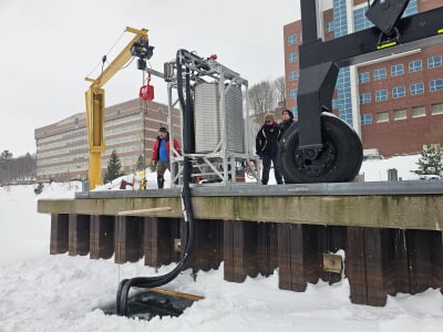 Three researchers stand on the docks above a frozen waterway, where insulated tubes are immersed in a large hole cut into the ice below.