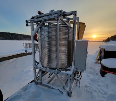 A large cylindrical tube encased in metal brackets rests on snow with pale yellow sun rising in the background.