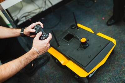 Hands holding a remote control above a flat, rectangular black and gold robot with antennae.