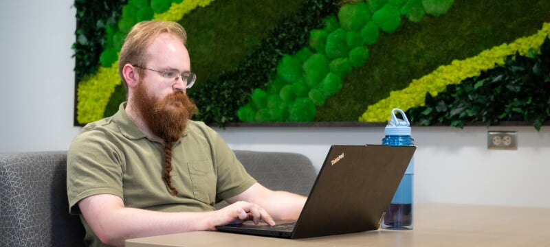 Elijah Nieman sits at a desk facing his laptop screen. A water bottle sits near him, and the wall behind him displays a large 3D art piece imitating layers of moss.