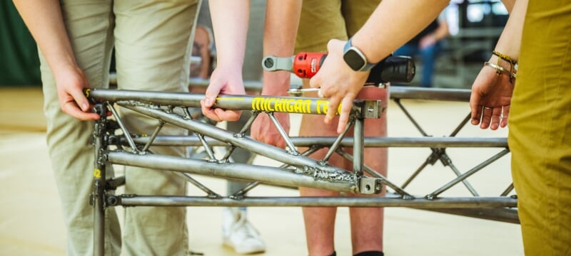 A close-up photo of steel bridge construction, with the words “Michigan Tech” visible along a steel beam among working hands.