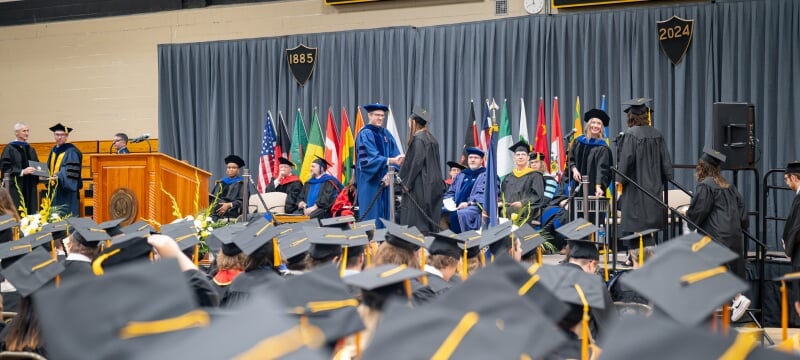 Students receiving diplomas on stage at 2024 Midyear Commencement, seen above the caps of those sitting down.