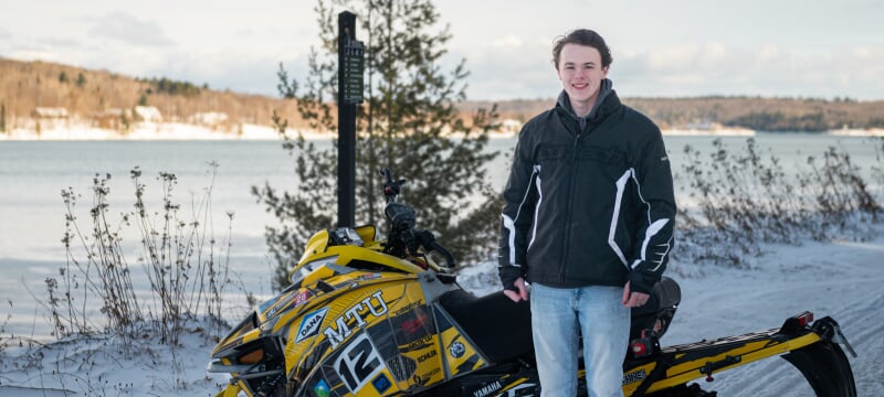Jared Ott stands in front of a yellow MTU snowmobile on a snowy day near Prince’s Point wearing a black jacket and blue jeans.