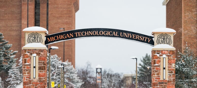 Archway with full univesity name in foreground with snow-covered trees and buildings in the background.