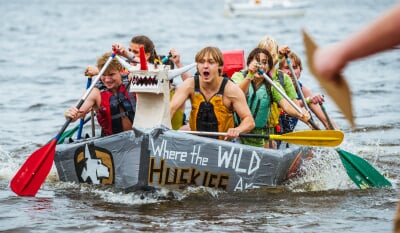 Eight college students in life jackets paddle a cardboard boat, decorated with Michigan Tech's Husky logo and the motto Where the Wild Huskies Are, through the water.