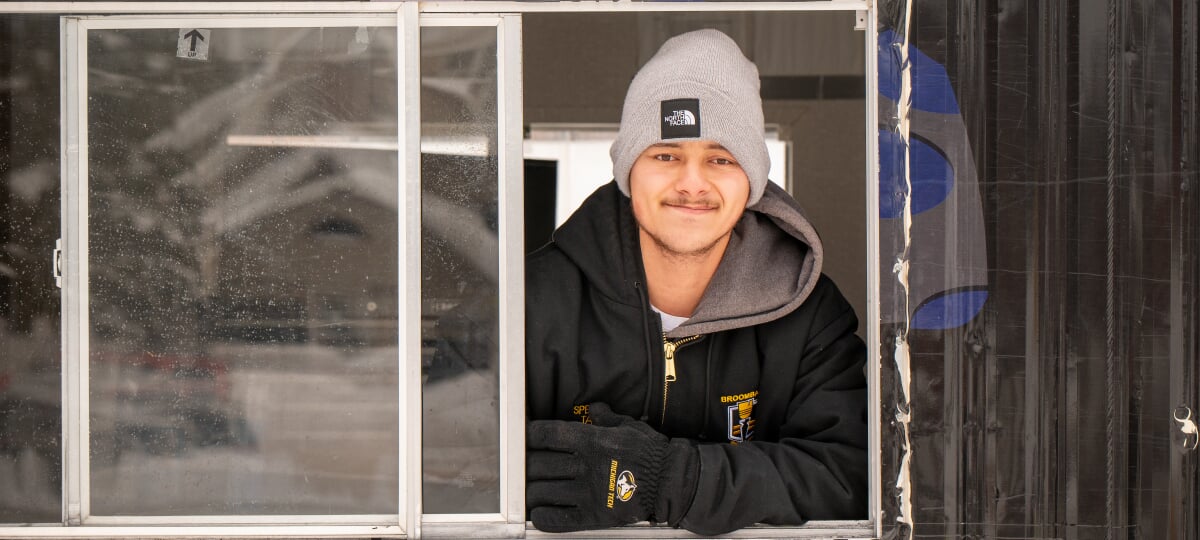 College of Business Husky Hieu Theriault puts his acumen to work on both broomball rinks and balance sheets. He’s shown here in Husky Nation’s iconic Cocoa Shack, where students can warm up with a hot beverage in between broomball games. Hieu Theriault looking out from the serving window of the cocoa shack trailer.